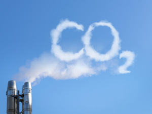 Ilustrační obrázek, který znázorňuje průmyslový komín, ze kterého stoupá kouř vytvářející nápis CO₂ na modré obloze.